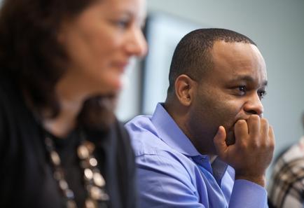 A man and a woman sitting in a conference room. The man is on the right side of the image with his chin resting on his hand. He is wearing a blue collared shirt and appears to be deep in thought. The woman on the left is slightly blurred but it seems like she is listening attentively to the man. She has shoulder-length dark hair and a necklace around her neck. The background is blurred so it is difficult to make out any other people in the room.