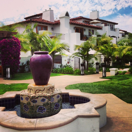 A beautiful courtyard with a large white building in the background. The building has a red tiled roof and is surrounded by palm trees and other tropical plants. In the foreground there is a circular fountain with a purple vase on top. The fountain is made of stone and has a floral design on it. The sky is blue and there are a few clouds in the sky. The courtyard is well-maintained with a variety of colorful flowers and plants.