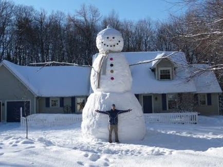 A large snowman made of snow in front of a house. The snowman is standing on its hind legs with its arms stretched out to the sides. It has a carrot nose two black eyes and a red button on its chest. The house in the background is a two-story building with a gray roof and white trim. The sky is blue and there are trees in the distance. The ground is covered in a thick layer of snow.