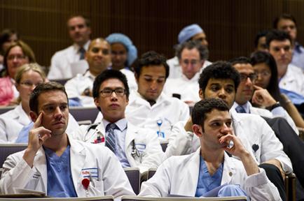 A group of medical professionals sitting in a lecture hall. They are all wearing white lab coats and blue scrubs. Some of them have stethoscopes around their necks indicating that they are medical professionals. The people in the image appear to be of different ages and genders and they are all looking towards the front of the room with serious expressions on their faces. The room has a wooden paneled wall in the background.