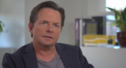 A close-up of a middle-aged man's face. He appears to be in his late 40s or early 50s with short light-colored hair and a serious expression. He is wearing a dark blue blazer and a gray t-shirt. The background is blurred but it seems to be an office or a living room with a plant and a bookshelf visible. The man is looking directly at the camera with a slight smile on his lips.