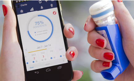 A person's hand holding a smartphone with a blue asthma inhaler in their other hand. The phone screen displays a dashboard with a percentage of 75% adherence and a date and time displayed on it. The person's nails are painted with red nail polish. The background is blurred but it appears to be an outdoor setting with greenery.