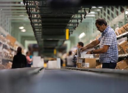 A man in a warehouse sorting through cardboard boxes on a conveyor belt. He is wearing a blue plaid shirt and glasses and is holding a cardboard box in his hands. He appears to be sorting through the boxes with a focused expression on his face. The warehouse is filled with shelves on both sides and there are other people in the background. The conveyor belts are lined up in rows and the ceiling is high and green. The image is taken from a low angle looking down on the man and the boxes.