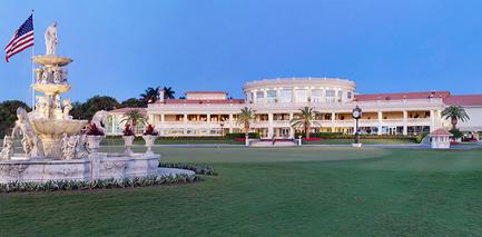A large luxurious building with a white exterior and a red roof. The building is surrounded by a well-manicured lawn and palm trees. In front of the building there is a large fountain with several statues and an American flag on top. The sky is blue and the sun is setting casting a warm glow over the scene. The overall atmosphere of the image is peaceful and serene.