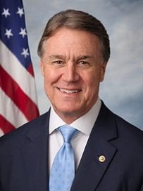A headshot of Joe Manchin a United States Senator from West Virginia (Democrat). He is wearing a navy suit with a small gold pin on the lapel a white dress shirt and a light blue tie. He is standing in front of an American flag. He is smiling and looking directly at the camera. He has short dark hair and appears to be in his late 60s or early 70s. The background is a plain blue sky with white clouds.