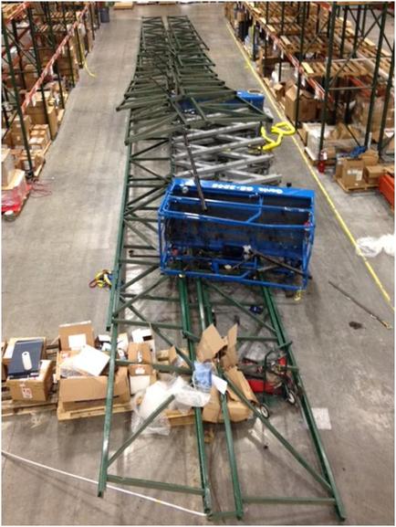 A large warehouse with rows of shelves on both sides. In the center of the image there is a long green metal structure that appears to be a conveyor belt or a hoist. The structure is made up of multiple trusses that are arranged in a grid-like pattern. On the right side of the structure there are several cardboard boxes and other items scattered on the floor. On top of the truss there appears to have a blue plastic container attached to it. The container is likely used for storing or transporting items. The warehouse is well-stocked with various tools and equipment.