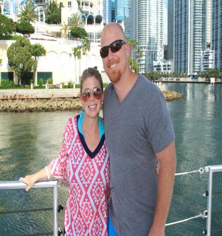 A man and a woman standing on a boat in a city. They are both smiling and posing for the camera. The man is bald and wearing sunglasses while the woman is wearing a pink and white patterned top and sunglasses. They appear to be happy and relaxed. In the background there are tall buildings and palm trees along the waterfront. The sky is blue and the water is calm.