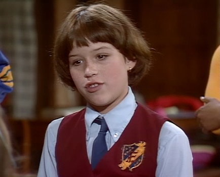 A close-up of a young boy's face. He appears to be in his late teens or early twenties. He has shoulder-length brown hair and is wearing a red vest with a gold emblem on the front. He is also wearing a blue collared shirt and a blue tie. The boy is looking directly at the camera with a serious expression on his face. In the background there are other people and a wooden wall.