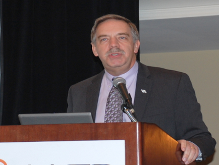A man standing behind a wooden podium with a microphone in front of him. He is wearing a dark suit and a purple tie. He has a mustache and is looking directly at the camera with a slight smile on his face. Behind him there is a black curtain and a white wall. On the podium there are two laptops. The man appears to be giving a speech or presentation.