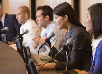 A group of four people sitting at a table with microphones in front of them. They appear to be engaged in a discussion or panel discussion. The table is covered with a white tablecloth and there are several computer monitors on it. The people are dressed in formal business attire and are seated in a row. The woman in the center of the image is speaking into one of the microphones while the other three people are listening attentively. The background is blurred but it appears to be a conference room with a large window and a curtain.