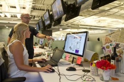 An office space with multiple computer monitors hanging from the ceiling. There are two people in the image a man and a woman sitting at a desk in front of a computer. The woman is typing on a laptop and the man is standing behind her pointing at the screen. The desk is cluttered with various items such as a vase of red flowers a bottle of wine and some papers. The room appears to be modern and well-lit.