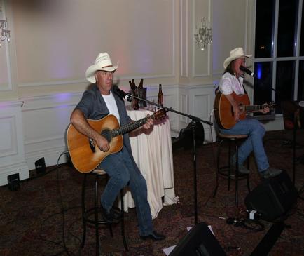 Two men sitting on stools in a room with white walls and a chandelier hanging from the ceiling. They are both wearing cowboy hats and playing acoustic guitars. The man on the left is playing an acoustic guitar and the man in the middle is singing into a microphone. There is a table in front of them with a white tablecloth and a vase of flowers on it. The room appears to be a banquet hall or event space.