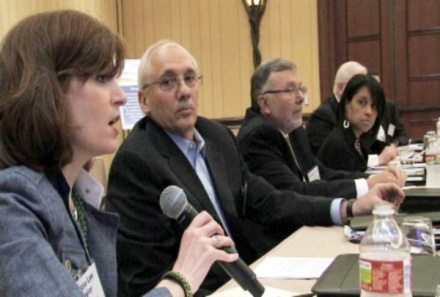 A group of people sitting around a table engaged in conversation. The room is well-lit and has wooden paneling on the walls. There are several microphones placed on the table and some water bottles can be seen nearby.