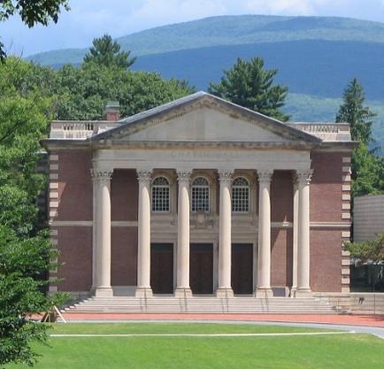 A large two-story brick building with a pediment on the top. The building has a flat roof and is surrounded by trees and greenery. In the background there is a mountain range with a clear blue sky. The front of the building has three columns with arched windows and a set of stairs leading up to the entrance. The ground is covered in green grass.