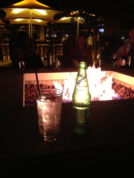 A table with a glass of water and a bottle of Sprite on it. The table is set up in front of a fire pit which is lit up with orange and yellow lights. There are several people sitting around the fire pit and umbrellas providing shade. The background is dark suggesting that the photo was taken at night.
