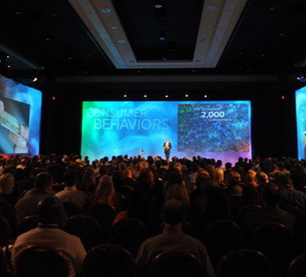 A large auditorium filled with people sitting in rows of chairs facing a stage. On the stage there is a man standing at a podium and giving a presentation. The stage is set up with two large screens on either side. The screens display the words "Consumer Behaviors" and "2000". The audience is attentively listening to the man's speech. The room is dimly lit with blue and red lights.