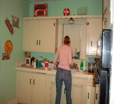 A woman standing in a kitchen. She is wearing a pink shirt and blue jeans and is standing in front of a white countertype with a sink. On the countertype there are various kitchen items such as a toaster a coffee maker a blender and a bottle of water. Above the sink there is a white cabinet with a red and white sign that reads "Coca-Cola." On the left side of the image on the right side there appears to be a refrigerator. The walls are painted in a light green color and there are several framed pictures hanging on the wall.
