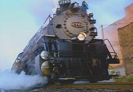 A steam locomotive numbered 2765 on a railway track. The locomotive is black in color and has a large round headlight in the center. The number 2765 is written in gold letters on the front of the locomotive. The train is emitting a plume of white smoke as it moves along the track. On the right side of the image there is a brick building and a power line. The sky is blue and the sun is setting casting a warm glow over the scene.