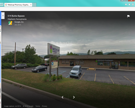 A street view of a building with a sign that reads "Medicare Pharmacy oliphant Pennsylvania". The building is a two-story structure with a brown exterior and a green roof. There are several windows on the front of the building and a parking lot in front of it. The sky is cloudy and there are trees and mountains in the background. There is a car parked in the lot and a few other cars parked nearby. The image appears to be taken from a Google Street View perspective.