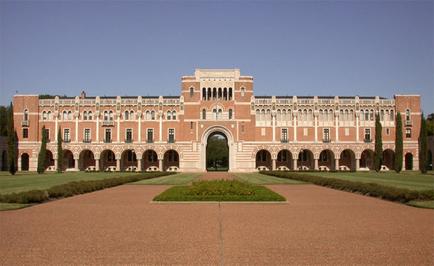 A large two-story brick building with a symmetrical entrance. The building has multiple levels with arched windows and columns and a large arched doorway in the center. The entrance is flanked by two smaller arches on either side. In front of the building there is a well-manicured lawn with a small garden in the foreground. The sky is blue and there are trees in the background. The ground is made of red bricks and the building appears to be well-maintained.