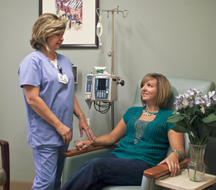 A nurse in a blue scrub suit standing next to a patient in a green armchair. The patient is sitting in the armchair and the nurse is standing on the left side of the image. The nurse is holding the patient's hand and appears to be explaining something to the patient. There is a medical equipment in the background including a blood pressure monitor and a vase of purple flowers on the table next to the chair. The wall behind the nurse has a framed picture hanging on it.