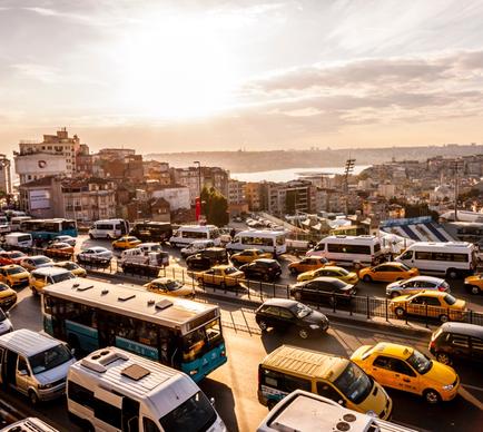An aerial view of a busy city street with multiple lanes of traffic. The street is filled with cars buses and taxis of different colors and models. The sky is orange and pink indicating that it is either sunrise or sunset. In the background there are several buildings and houses and a body of water can be seen in the distance. The overall mood of the image is bustling and bustling.