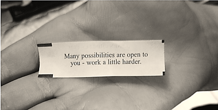 A black and white photograph of a person's hand holding a small piece of paper with a quote written on it. The quote reads "Many possibilities are open to you - work a little harder." The paper appears to be slightly crumpled and has a torn edge. The background is blurred so the focus is on the hand and the quote.