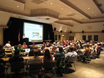 A large conference room with a high ceiling and rows of chairs. The room is filled with people sitting at long tables facing a stage where a man is giving a presentation. The man is standing at a podium and appears to be giving a speech. On the stage there is a large screen with an image of a map and some text. There are two flags on either side of the screen one is the American flag and the other is the United States flag. The walls of the room are decorated with the Olympic rings and the words "USA" and "Olympic Games." The people in the room appear to be engaged in the presentation.