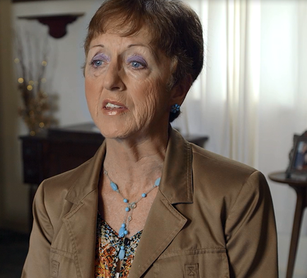 A close-up of an elderly woman's face. She appears to be in her late 60s or early 70s with short dark hair and blue eyes. She has a serious expression on her face and is looking off to the side with a slight smile. She is wearing a beige blazer over a colorful blouse with a leopard print pattern and a blue necklace. The background is blurred but it seems to be a living room with a window and a vase of flowers.