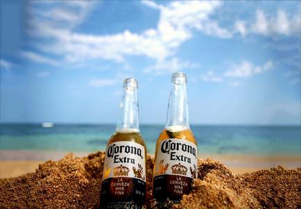 Two Corona Extra beer bottles lying on top of a pile of sand on a beach. The bottles are filled with a golden-colored beer and have the Corona Extra logo on the front. The background shows a beautiful blue sky with white clouds and the ocean in the distance. The sand is a light brown color and appears to be freshly sanded. The overall mood of the image is peaceful and serene.