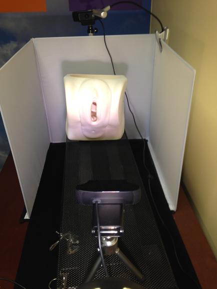 A white cube-shaped object on a black mat in a room with a yellow wall in the background. The cube appears to be made of a soft plush material and has a small hole in the center possibly for ventilation. The object is connected to a tripod with a camera attached to it. The tripod is positioned in front of the cube and there is a small black object on the mat next to it possibly a microphone or a speaker.