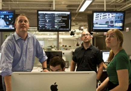 A group of four people in an office setting. There are three people in the foreground two men and two women standing in front of a large computer monitor. The man on the left is wearing a blue shirt and is looking up at the screen with a serious expression on his face. The woman on the right is sitting at a desk with a laptop in front her. In the background there are multiple computer monitors displaying various images and text. The room appears to be modern and well-lit with fluorescent lights.