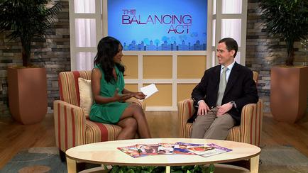 A man and a woman sitting in a TV studio. They are facing each other and appear to be engaged in a conversation. The woman is wearing a green dress and is holding a piece of paper in her hand. The man is also wearing a suit and tie. They both have serious expressions on their faces. In front of them there is a round wooden coffee table with a magazine on it. The background of the image is a TV screen with the words "The Balancing Act" and a city skyline. There are two potted plants on either side of the screen.