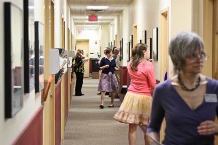 A long hallway with a group of people walking down it. On the left side of the hallway there are rows of framed pictures hanging on the wall. The walls are painted in a light beige color and there is a red exit sign at the end of the corridor. The people in the hallway appear to be of different ages and genders and they are walking in different directions. Some of them are wearing casual clothes while others are dressed in more formal attire. The hallway appears to be well-lit and has a carpeted floor.