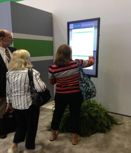 A group of three people standing in front of a large screen in an exhibition hall. The screen is displaying a presentation slide with a blue background and white text. The slide appears to be a presentation on a computer screen. The people in the image are looking at the screen intently and appear to be engaged in the presentation. On the left side of the screen there is a man wearing a suit and tie and on the right side there are two women one wearing a striped shirt and the other wearing a black and white striped shirt. The woman in the striped shirt is pointing to the screen with her hand while the other two women are standing next to her. There is a plant in the background and the floor is made of concrete.