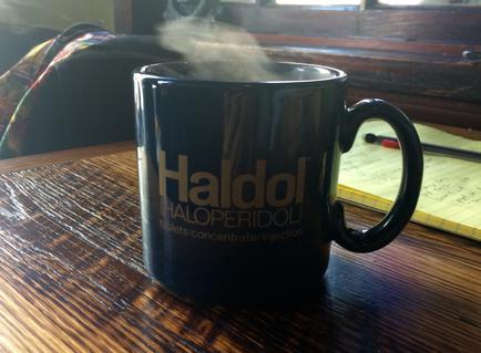 A black coffee mug with the word "aldol" written in white on it. The mug is sitting on a wooden table with a notebook and a pen next to it. There is a steam rising from the mug indicating that it is hot or steaming. The background is blurred but it appears to be a room with a window and a plant.