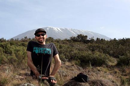 A man standing on a rocky hilltop with a mountain in the background. He is wearing a black t-shirt with the words "Redefine Possible" printed on it a black baseball cap and sunglasses. He has a backpack on the ground next to him and is smiling at the camera. The mountain appears to be covered in snow and there are trees and shrubs scattered around. The sky is blue and the overall scene is peaceful and serene.