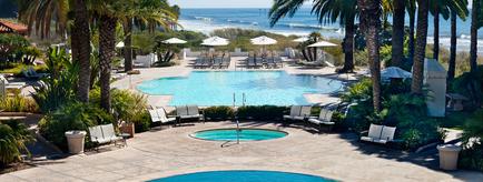 A large swimming pool surrounded by palm trees and other tropical plants. The pool is rectangular in shape and has a hot tub in the center. There are several lounge chairs and umbrellas scattered around the pool providing a perfect spot to relax and enjoy the view of the ocean. The sky is blue and the ocean can be seen in the background. The overall atmosphere of the image is peaceful and relaxing.