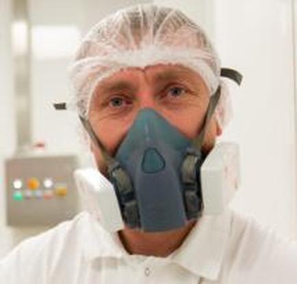 A man wearing a full face respirator mask. He is wearing a white lab coat and a white hairnet on his head. The mask is blue in color and has a filter attached to it. The man is looking directly at the camera with a serious expression on his face. In the background there is a control panel with various buttons and switches. The image appears to be taken in a laboratory or laboratory setting.