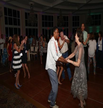 A group of people dancing in a large room with a high ceiling and chandeliers. The room appears to be a ballroom or banquet hall as there are large windows on the walls and a wooden floor. The people in the image are of different ages and genders and they are all dressed in formal attire. In the center of the image there is a man and a woman holding hands and dancing together. The man is wearing a white shirt and blue jeans and the woman has long dark hair. They are both smiling and appear to be enjoying themselves. There are other people around them some of whom are clapping and cheering. Some of them are wearing hats and dresses while others are wearing casual clothes. The overall atmosphere is lively and festive.