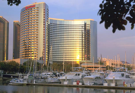 A marina with several boats docked in the water. In the center of the image there is a tall modern building with a curved glass facade. The building has a red and white logo on the top left corner. The sky is blue and the sun is setting in the background casting a warm glow over the scene. The marina is surrounded by trees and other buildings creating a peaceful atmosphere.