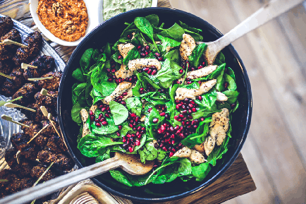 A large black bowl filled with a salad on a wooden table. The salad is made up of fresh green spinach leaves topped with sliced chicken and pomegranate seeds. There are two wooden spoons in the bowl ready to be used to mix the ingredients together. On the left side of the table there is a plate of grilled meat skewers and on the right side there are two small bowls of hummus and a small bowl of pesto sauce. The table is decorated with a woven basket and a woven placeman.