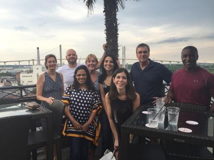 A group of nine people posing for a photo on a rooftop terrace. There are nine people in the photo nine women and one man all of whom are smiling and looking at the camera. They are standing in front of a palm tree and there are two tables and two chairs set up on the terrace with drinks on them. In the background there is a view of a city skyline with buildings and a bridge. The sky is cloudy and the overall mood of the photo is happy and relaxed.