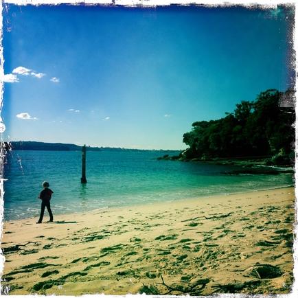 A photograph of a beach with a clear blue sky and the ocean in the background. The beach is sandy and the water is a beautiful turquoise color. On the right side of the image there is a small island with trees and bushes on the shore. In the distance there are a few people walking along the beach. The sky is a bright blue with a few white clouds scattered across it. The overall mood of the photograph is peaceful and serene.