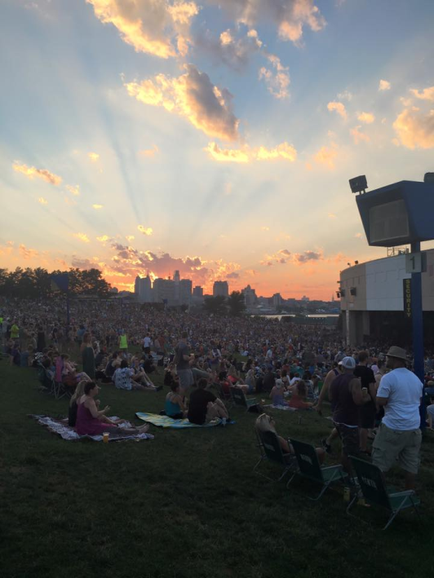 A large crowd of people gathered on a grassy field at a music festival. The sky is filled with orange and yellow hues indicating that it is either sunrise or sunset. The sun is shining brightly creating rays of light that are shining down on the crowd. In the background there are buildings and trees suggesting that the festival is taking place in a city. The people in the crowd are sitting on lawn chairs and some are standing enjoying the music. The overall mood of the image is lively and energetic.