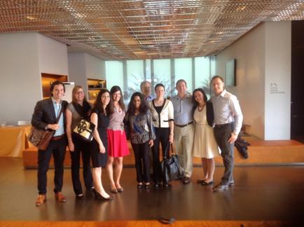A group of nine people standing in a large room with a high ceiling and a large window in the background. They are all smiling and appear to be posing for a photo. The room has a wooden floor and white walls and there is a wooden bench on the right side of the image. The people are dressed in formal attire with some wearing suits and others wearing dresses. There are nine people in total and they are all looking at the camera.