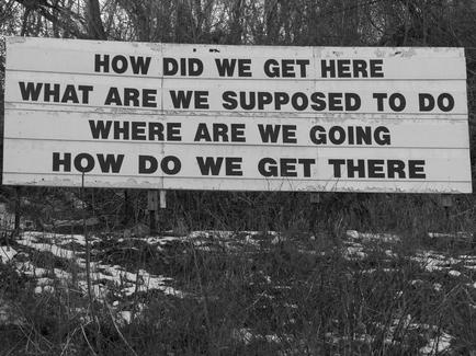 A black and white photograph of a large white sign with black text on it. The sign is located in a wooded area with trees and bushes in the background. The text on the sign reads "How did we get here what are we supposed to do where are we going how do we get there?" The sign appears to be old and weathered with some of the text faded and discolored. The sky is overcast and the ground is covered in snow.