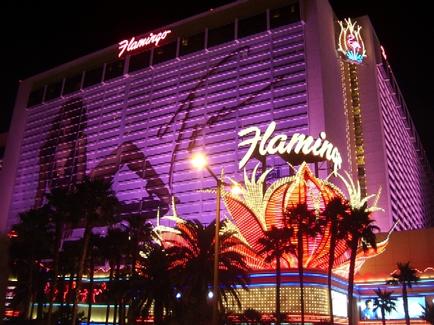 The exterior of the Flamingo Hotel and Casino in Las Vegas Nevada. The hotel is a large modern building with a curved facade and multiple levels. The building is lit up with colorful lights creating a vibrant and eye-catching display. The word "Flamingo" is prominently displayed on the top of the building in large bold letters. In front of the hotel there are palm trees and a large Ferris wheel-like structure which is also lit up in bright colors. The sky is dark making the colors of the lights stand out even more.