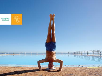 A man performing a handstand on the edge of a swimming pool. He is wearing blue shorts and is upside down with his arms and legs stretched out in front of him. His head is tilted back and his eyes are closed as if he is enjoying the moment. The pool is surrounded by a clear blue sky and there are a few people visible in the background. On the top left corner of the image there is a logo that reads "Live your life".