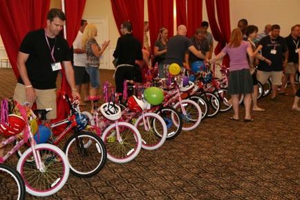 A group of people gathered in a room with red curtains in the background. In the center of the image there are several pink bicycles lined up in a row. Each bicycle has a colorful helmet attached to the front wheel. The people in the image appear to be of different ages and genders and they are all looking at the bicycles with interest. Some of them are wearing ID cards suggesting that they are at an event or gathering.